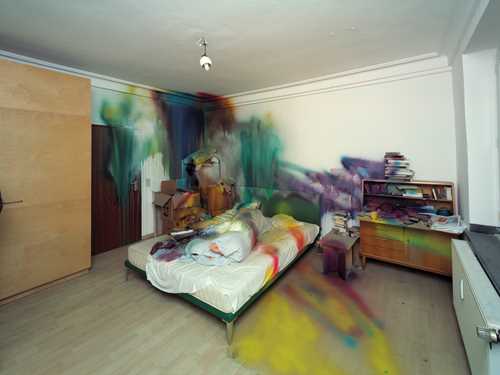 A room with a bed in the centre and various pieces of furniture on either side, all partially spray-painted in different colours. Photograph.