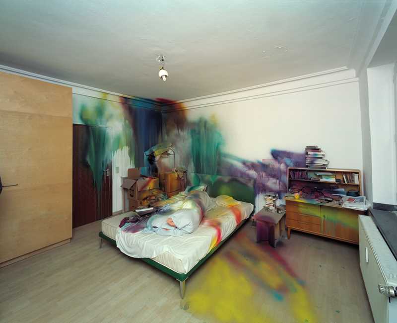 A room with a bed in the centre and various pieces of furniture on either side, all partially spray-painted in different colours. Photograph.