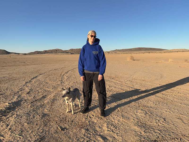 Vi ser en person i helfigur med blå hettegenser, svarte bukser og solbriller stående i et ørkenlandskap med en hund ved siden av seg. I bakgrunnen er det fjell og himmelen er knallblå. Fotografi