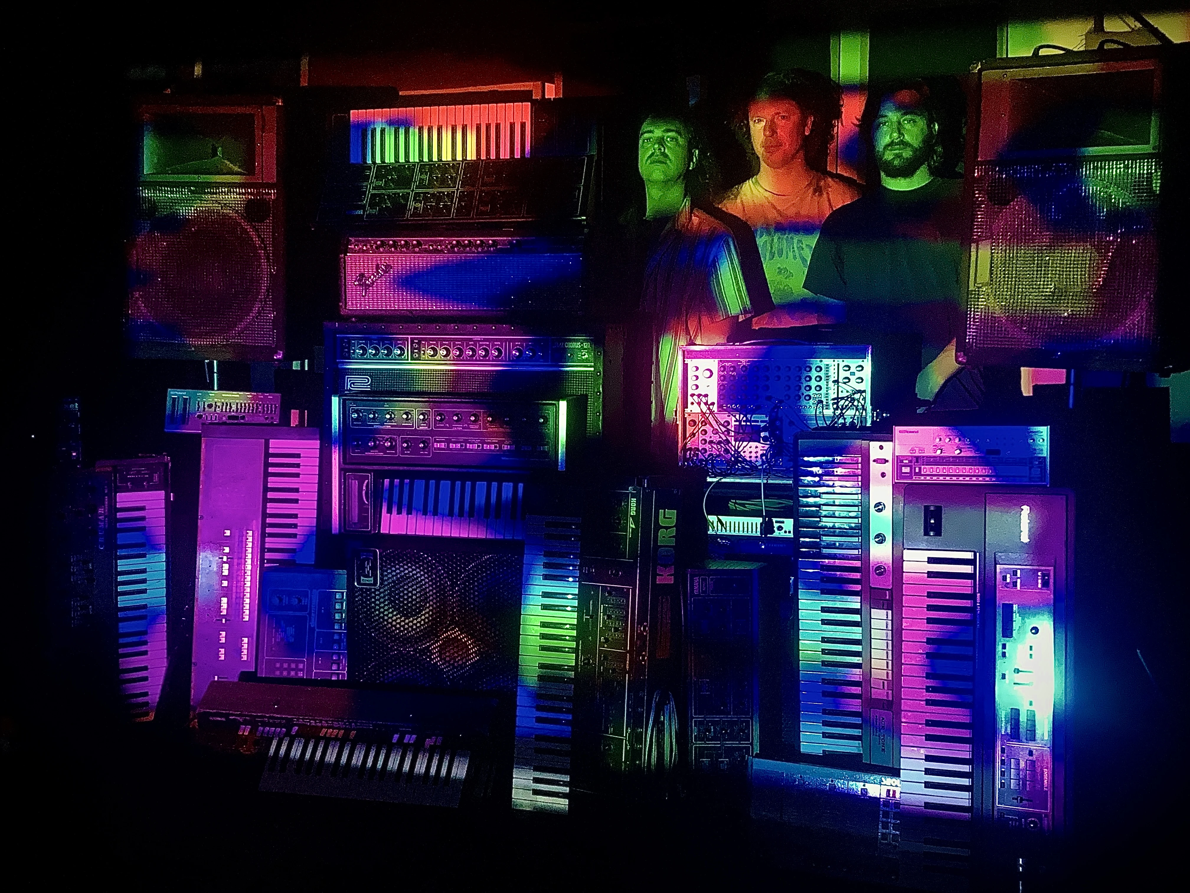 Bilde av en stabel med synthesizere og tre menn som står bak
