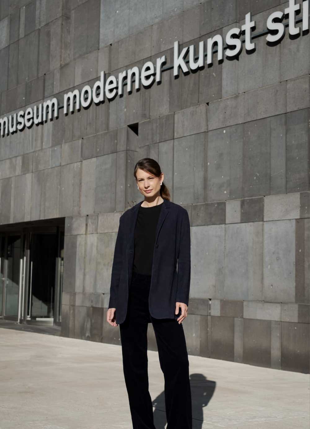Fatima Hellberg stands outside the Museum moderner Kunst Stiftung Ludwig in Vienna. She is shown full-length, wearing black trousers and a sweater with a dark blue blazer. Photograph.