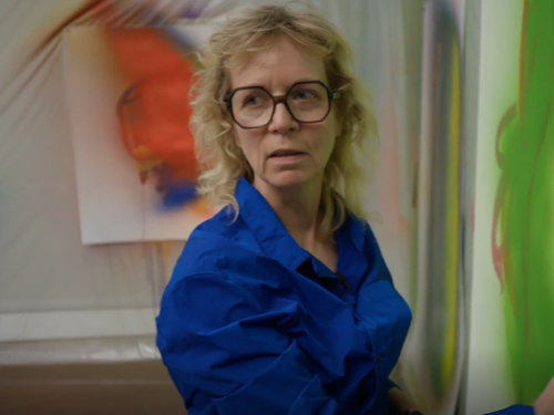 Portrait of a blond woman with big glasses and a blue shirt holding a green painting