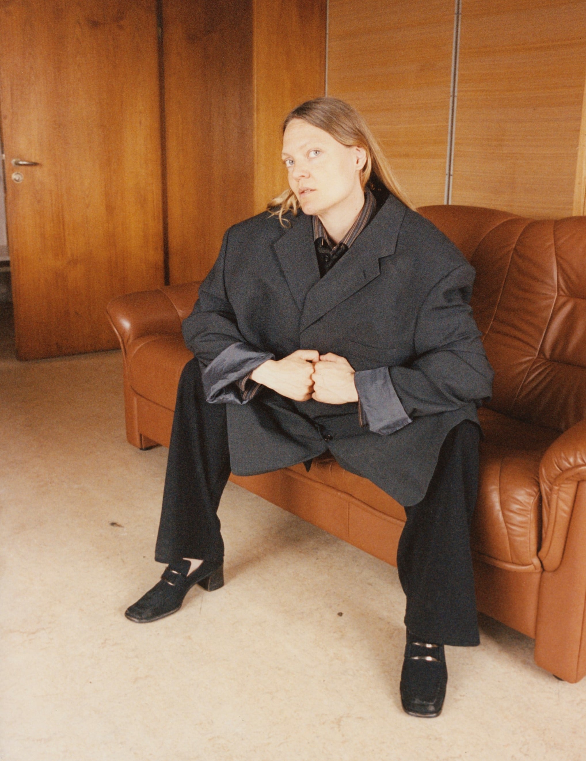 A woman sits on a brown leather sofa. She is wearing a large grey blazer, black trousers, and black shoes with square toes, a buckle, and high heels. Photograph.