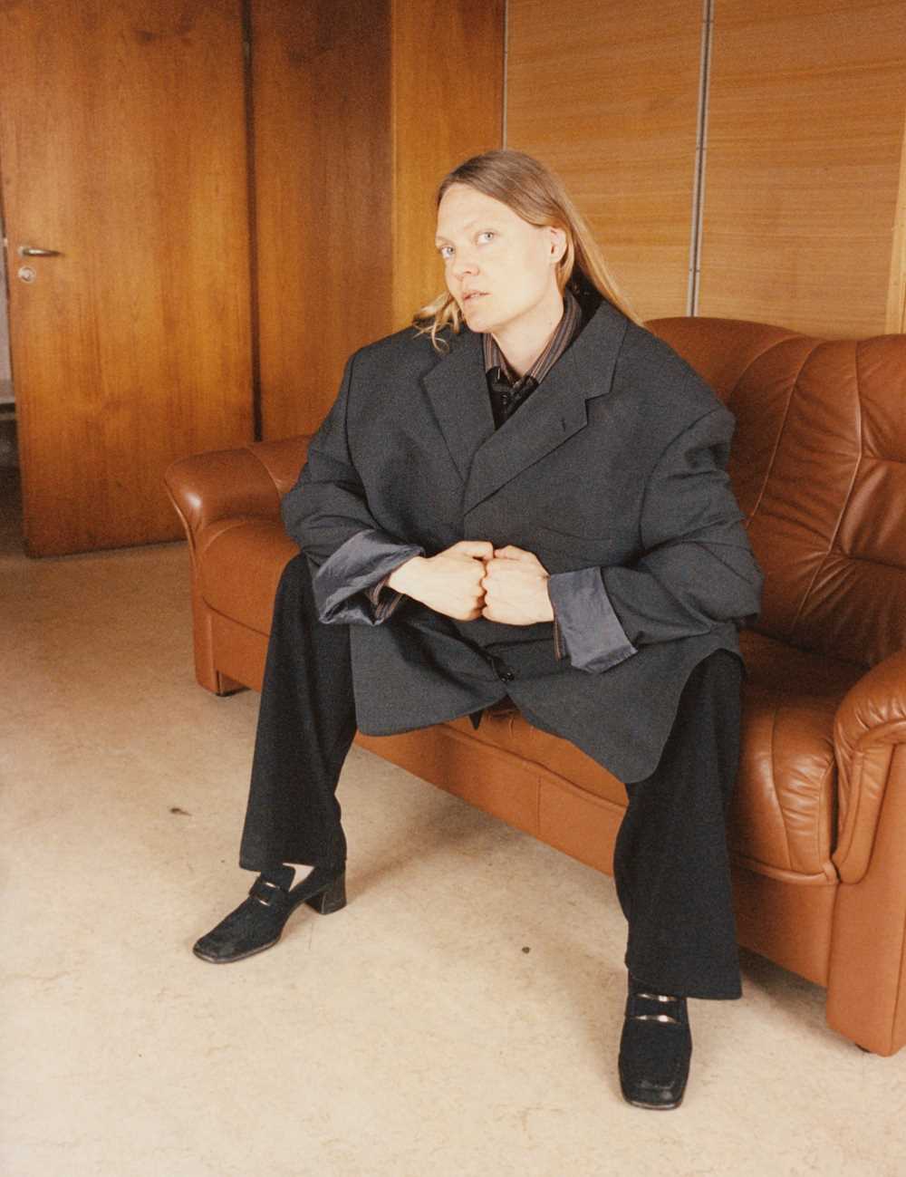 A woman sits on a brown leather sofa. She is wearing a large grey blazer, black trousers, and black shoes with square toes, a buckle, and high heels. Photograph.