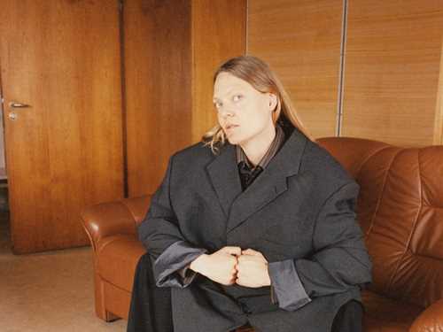 A woman sits on a brown leather sofa. She is wearing a large grey blazer, black trousers, and black shoes with square toes, a buckle, and high heels. Photograph.