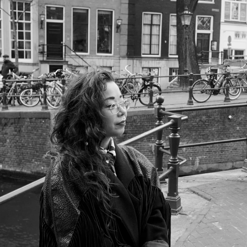 A black-and-white photo of a woman with long, wavy hair and glasses. She wears a leather jacket and is looking to the right side of the image frame with a calm expression. A city setting appears in the background.