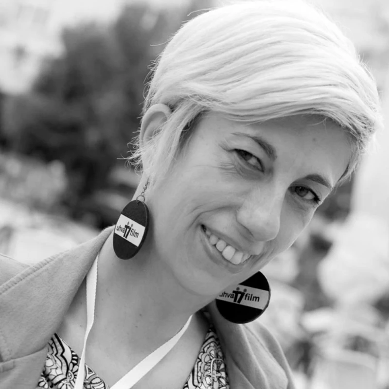 A black-and-white photo of a smiling woman with short hair, wearing a blazer and blouse, with large black earrings and a white ribbon around her neck. A city setting appears in the background.