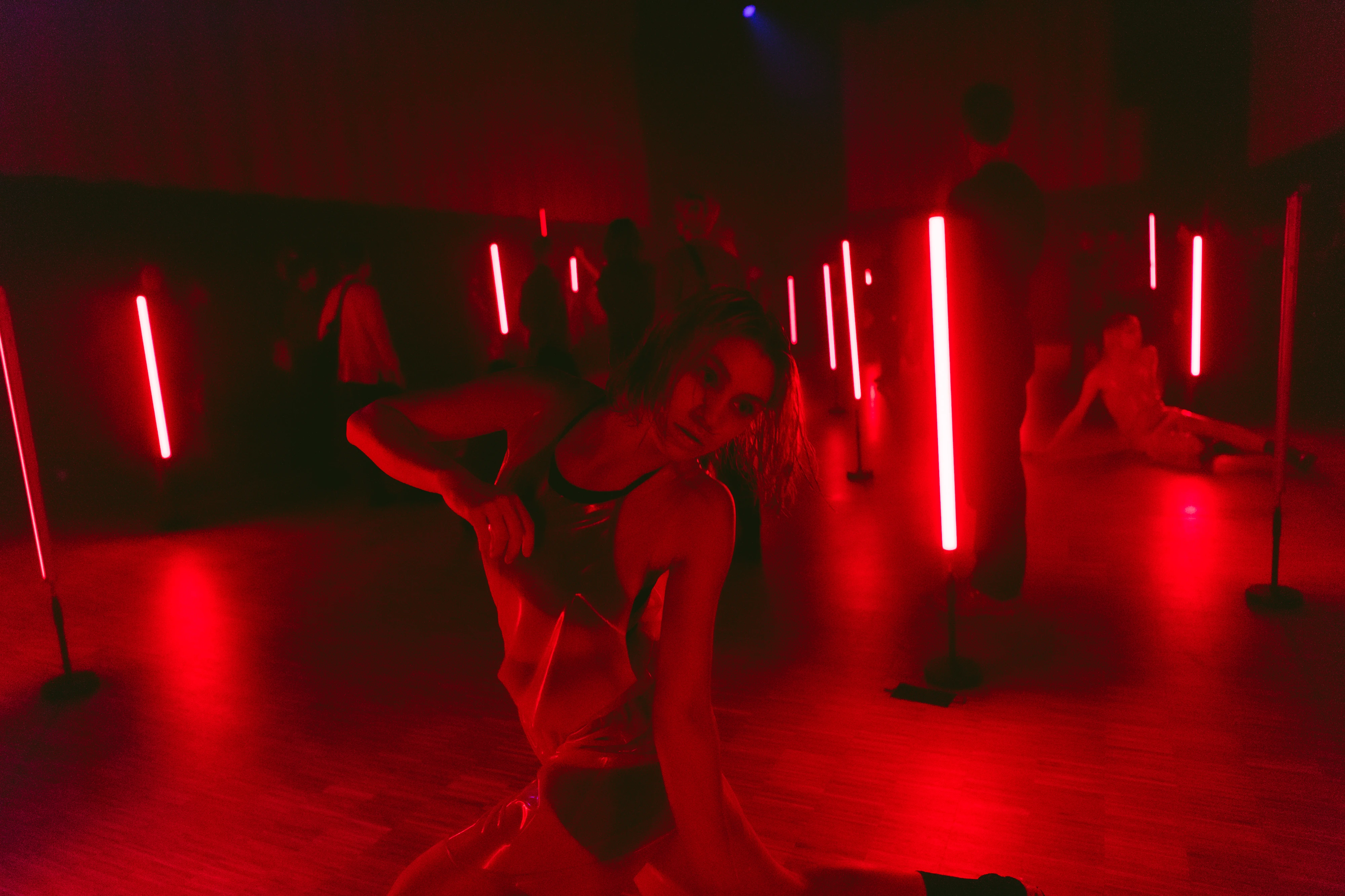 People wearing transparent plastic clothing move around a dark room illuminated by red fluorescent lights.