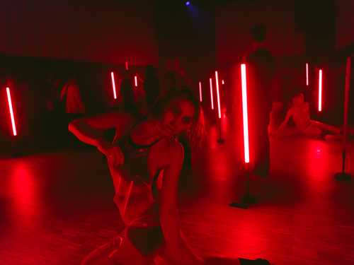 People wearing transparent plastic clothing move around a dark room illuminated by red fluorescent lights.