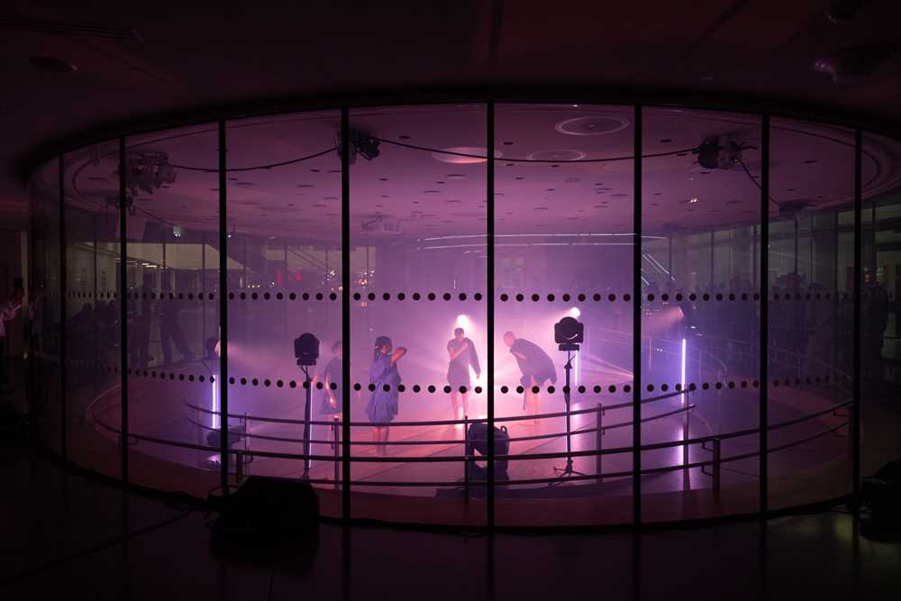 Four people dance inside a glass room lit by spotlights with purple light.