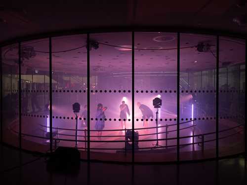 Four people dance inside a glass room lit by spotlights with purple light.