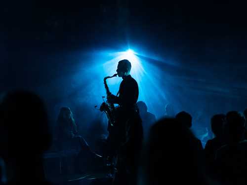 Saxophonist Bendik Giske stands and plays in the middle of a crowd seated on the floor, listening. The room is dark, illuminated by blue light. Photograph.