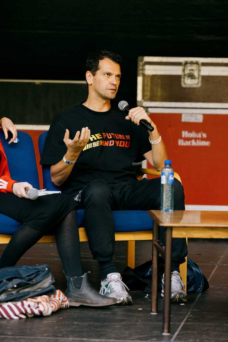 Performance artist Manuel Pelmuș sits on stage, speaking into a handheld microphone. Photograph.