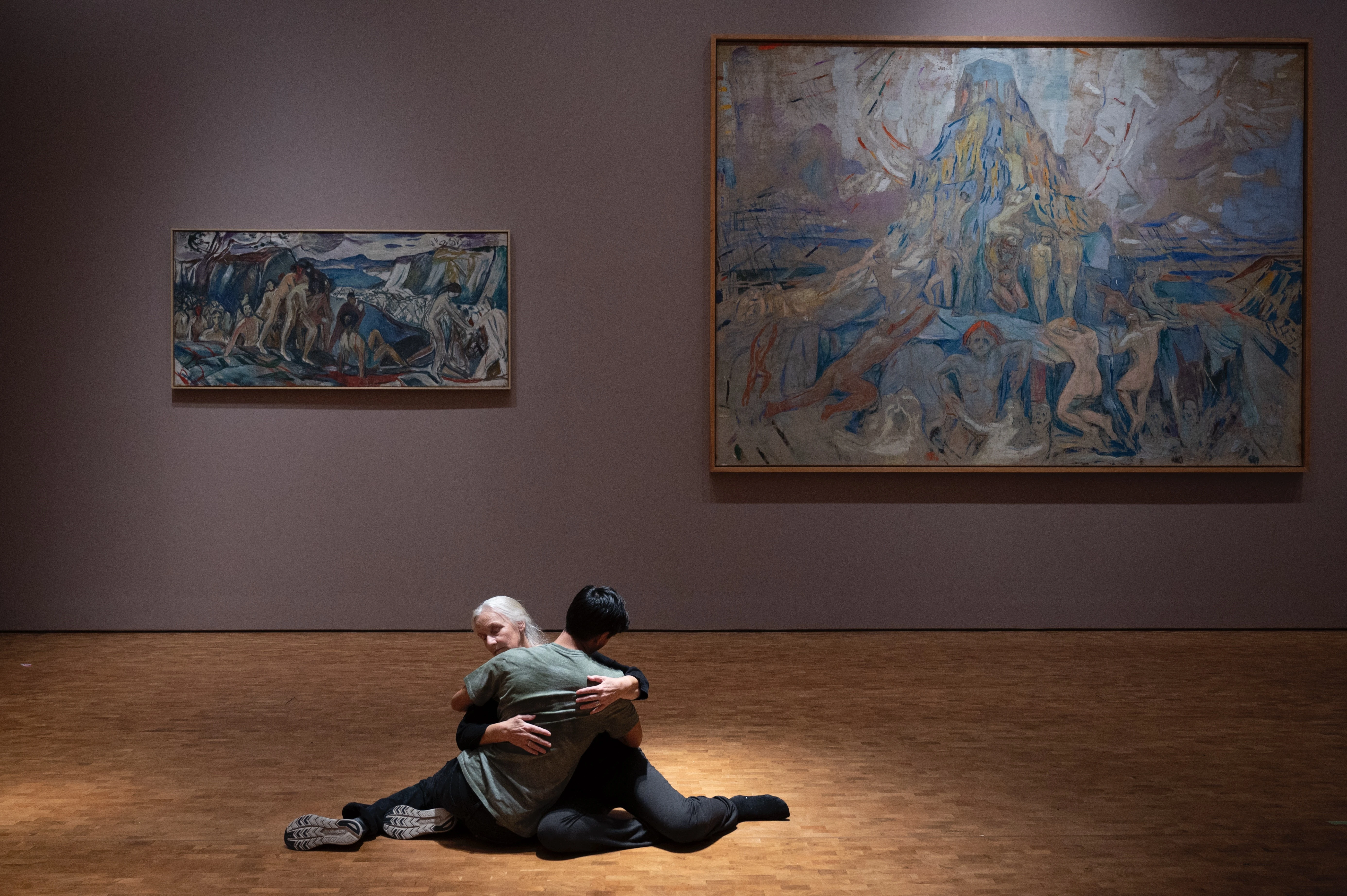 Two people sit on the floor in an embrace. In the background is Edvard Munch's painting The Human Mountain. Photograph.