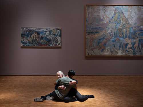 Two people sit on the floor in an embrace. In the background is Edvard Munch's painting The Human Mountain. Photograph.