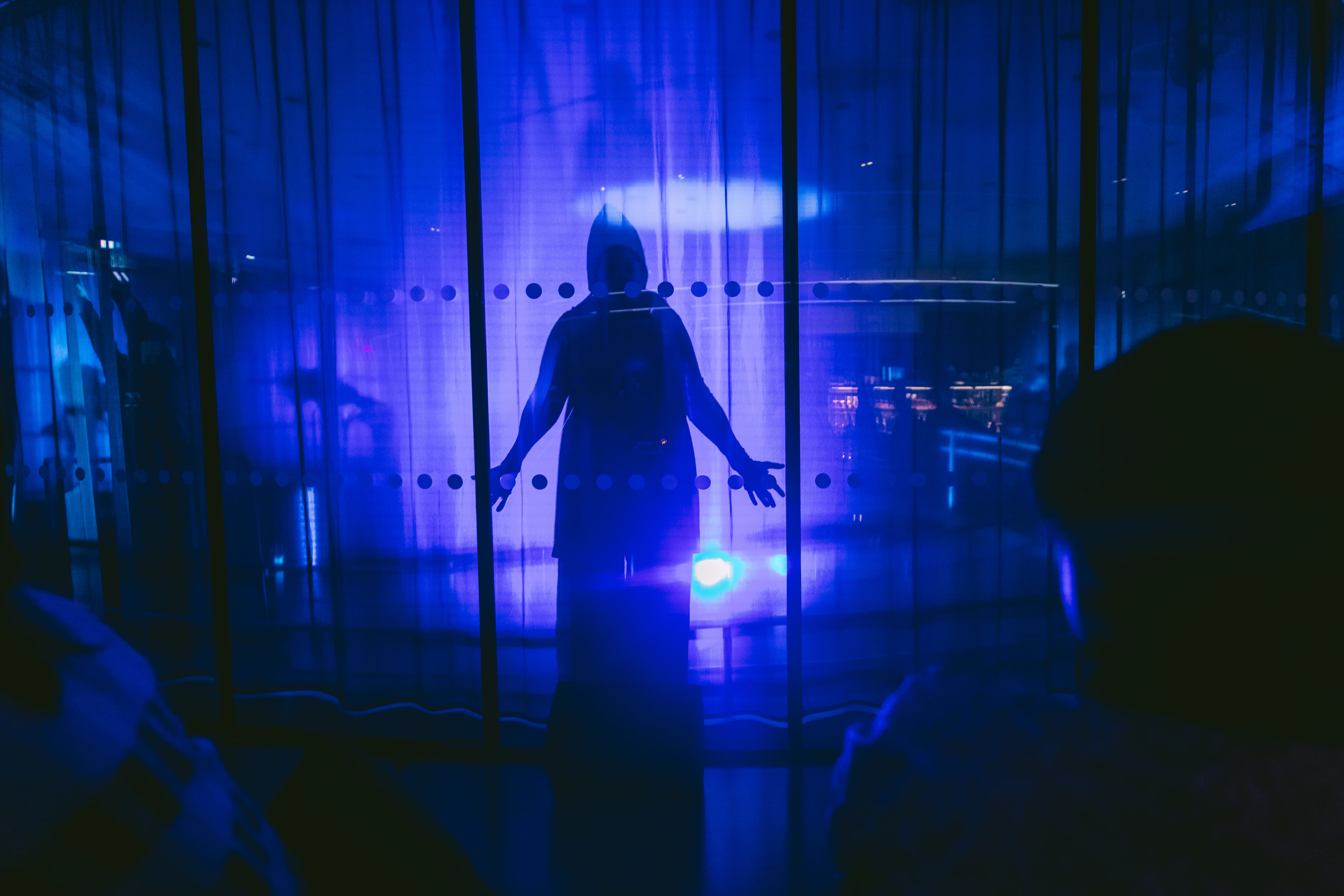 A cloaked figure stands behind a glass wall in a room illuminated by blue light. Photograph.