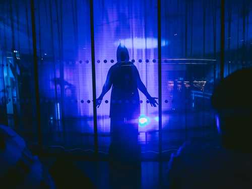 A cloaked figure stands behind a glass wall in a room illuminated by blue light. Photograph.