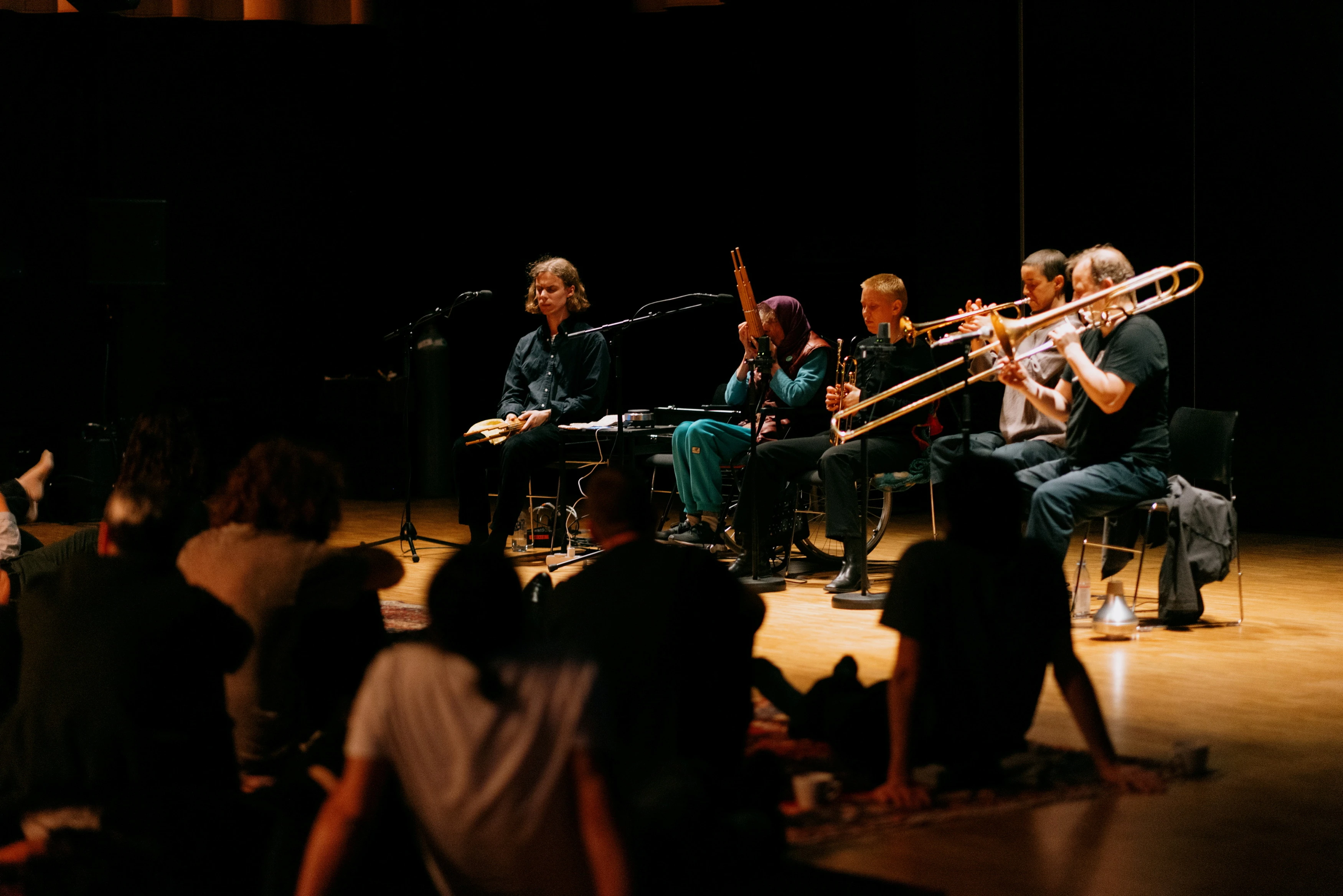 Five people with different instruments sit on individual chairs, performing in front of an audience seated on blankets on the floor. Photograph.