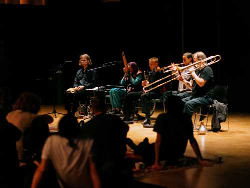 Five people with different instruments sit on individual chairs, performing in front of an audience seated on blankets on the floor. Photograph.